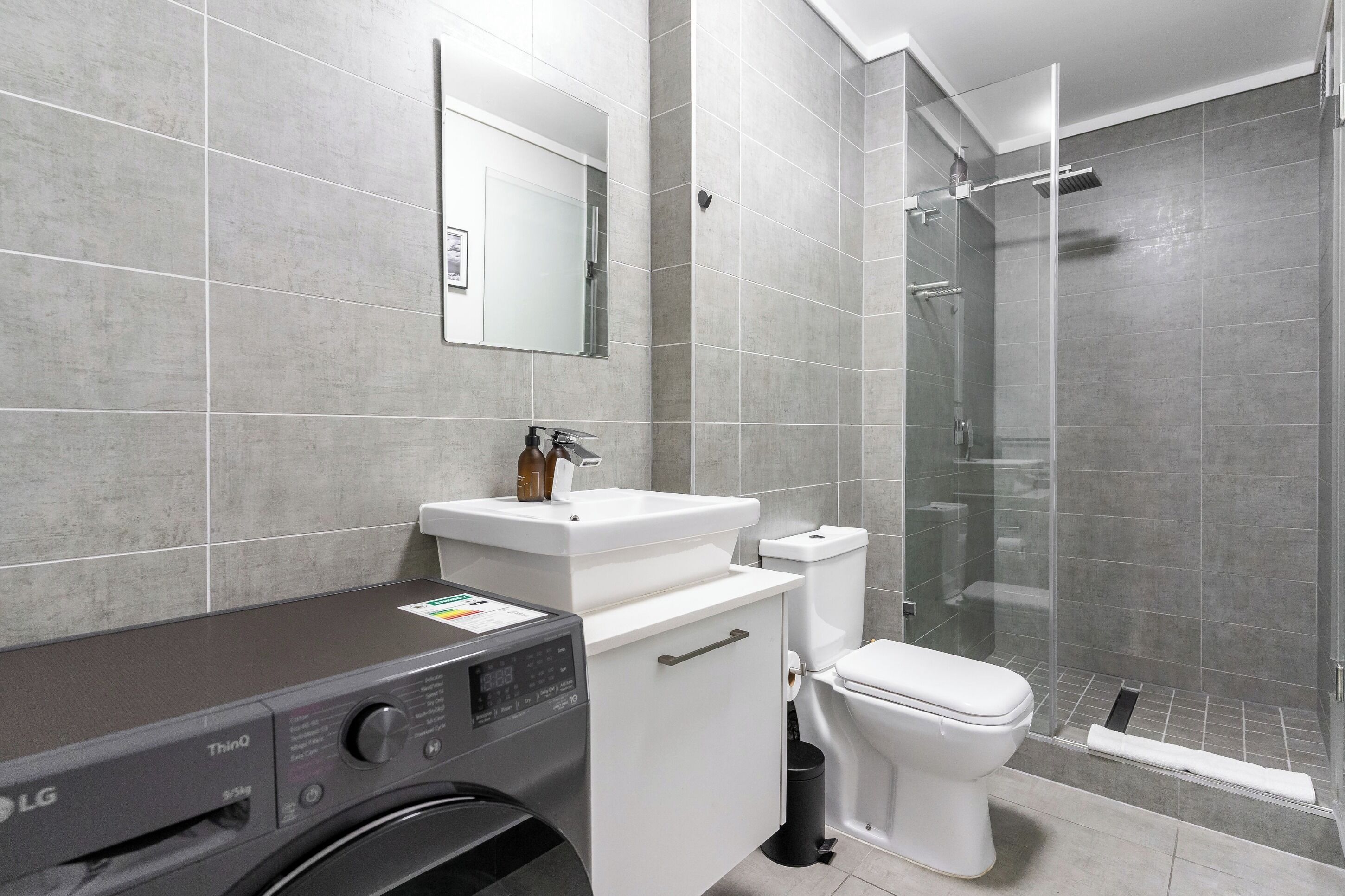Een moderne badkamer met een grijze betegelde muur, een wit wastafelmeubel, een toilet, een douche met glazen wand en een wasmachine.