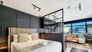 Ein modernes Hotelzimmer mit einem großen Bett, einer Sitzecke mit einer Couch und Sesseln sowie Blick auf die Stadt durch große Fenster.