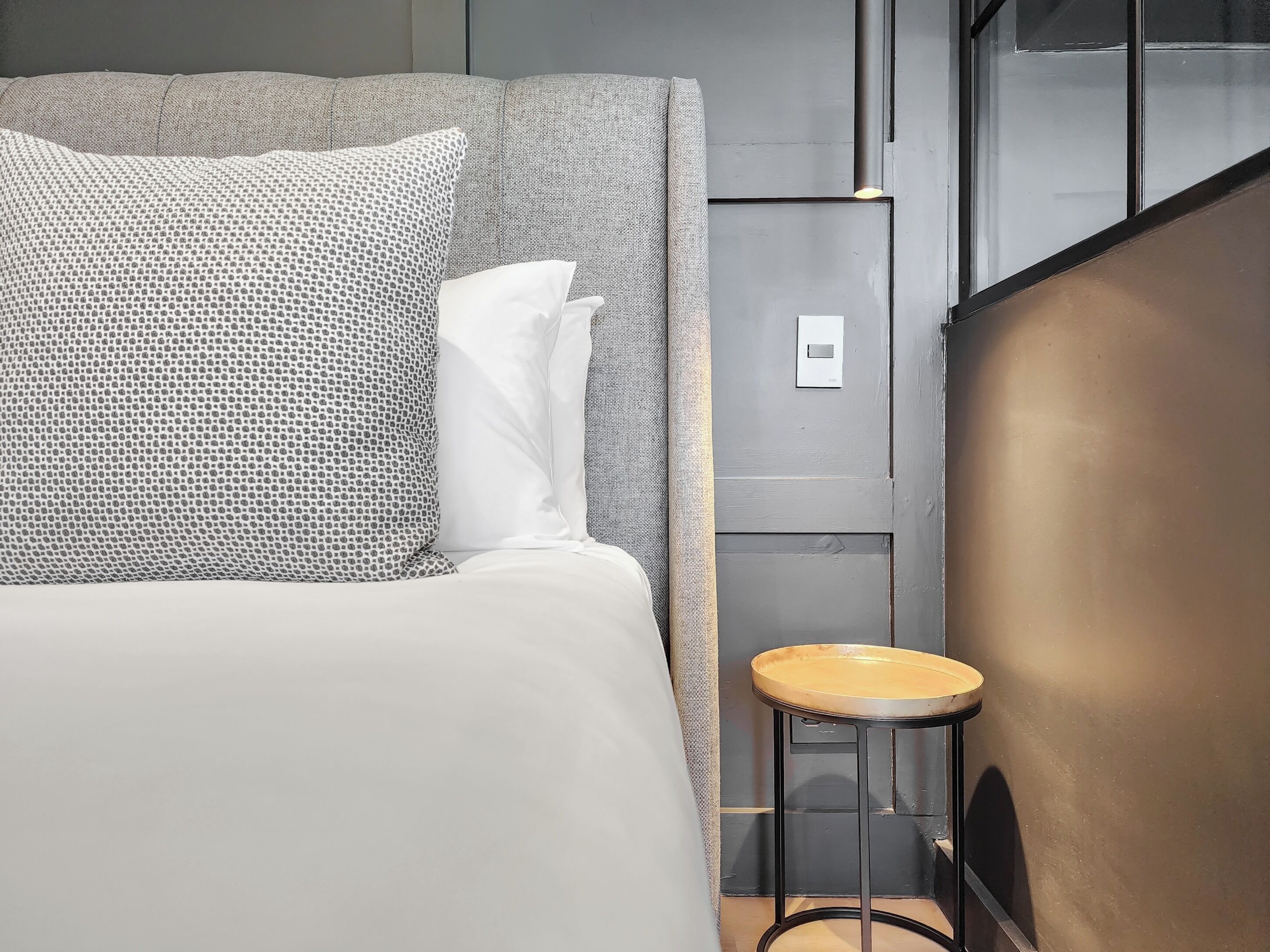 Quarto de hotel moderno com uma cama grande, um banquinho pequeno e um candeeiro fixado na parede.
