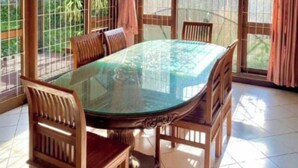 A round glass-top dining table with wooden chairs, surrounded by large windows with curtains. - Hotel O Bougenville Resort (Cipanas)