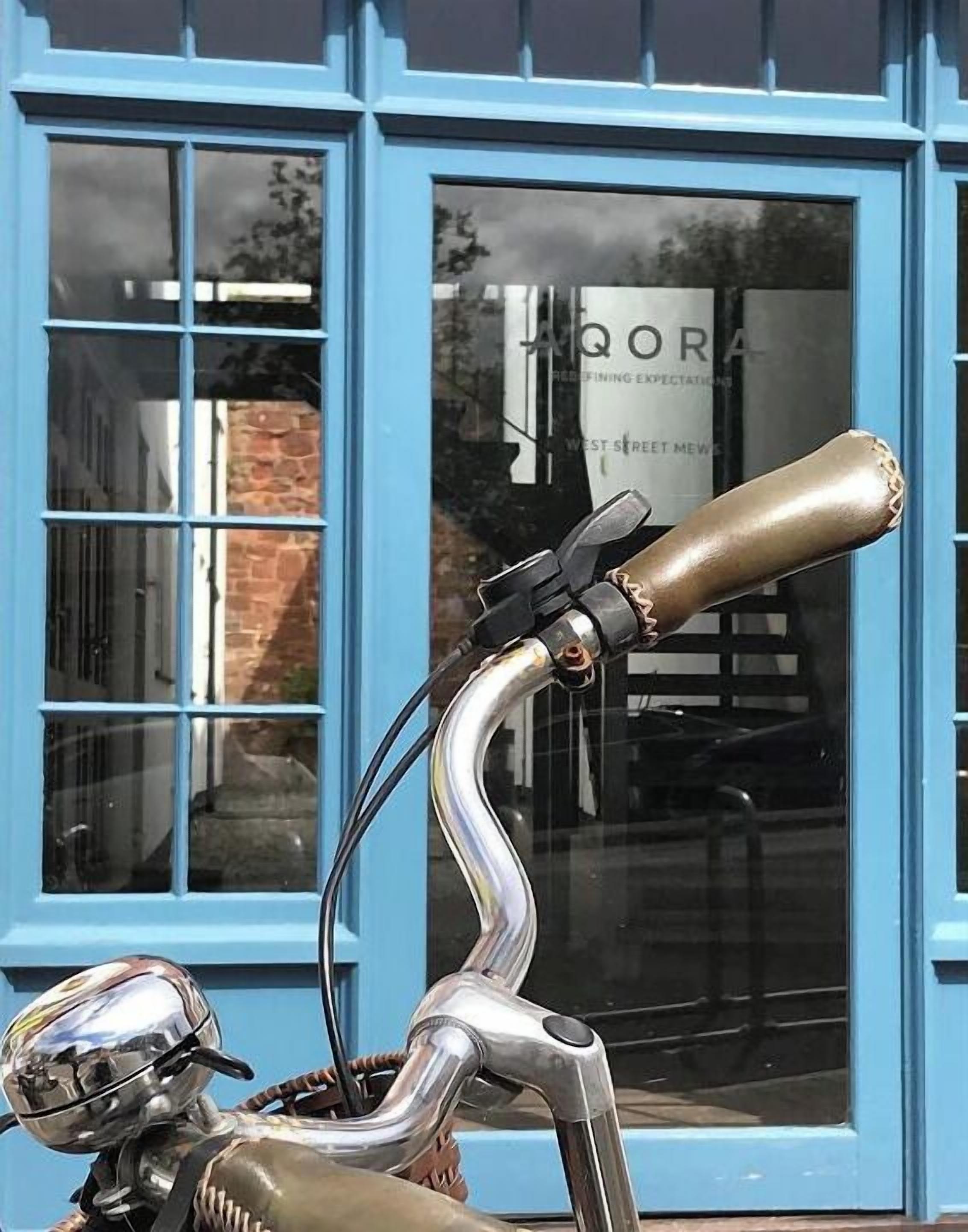 A bicycle with a unique handlebar design and a reflective headlight, parked in front of a building with blue trim and a window displaying the text "AQORA".