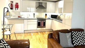 A modern kitchen with white cabinets, a microwave, and a stove. There is a dining area with a glass table and chairs, and a brown leather sofa with patterned cushions. - West Street Mews Serviced Accommodation (Exeter)