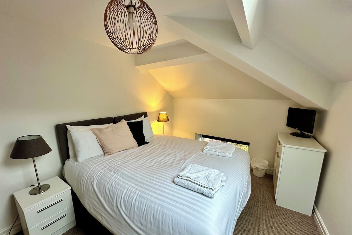 Une chambre bien rangée, avec un lit, une lampe de chevet et un petit bureau équipé d’un écran.