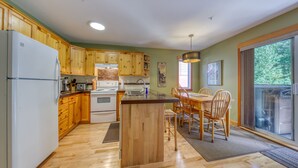 A kitchen with wooden cabinets, a central island, a dining table with chairs, and a large window. - Ski-in Townhome - Private Hot Tub - Fernie Resort (Fernie)