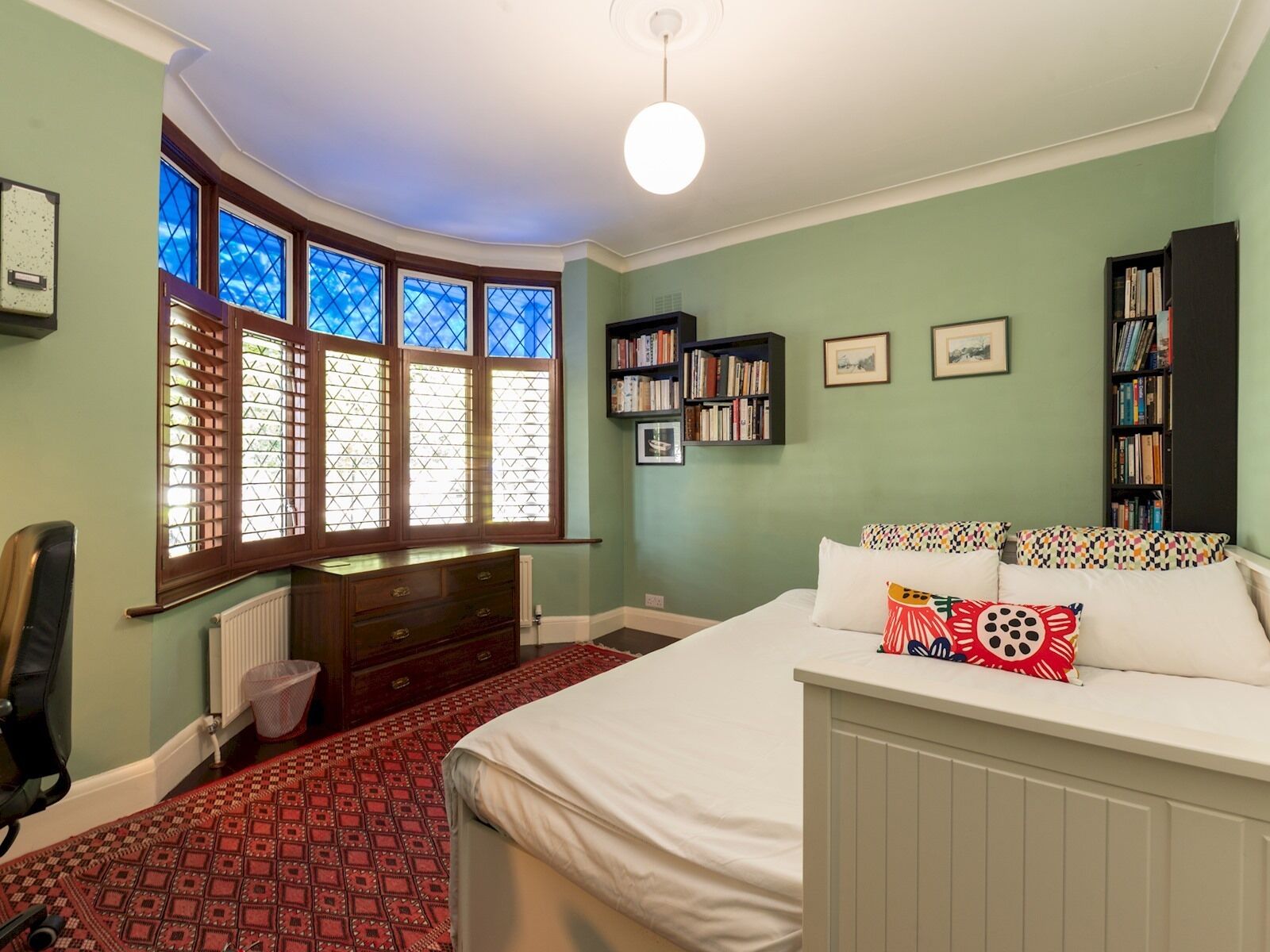 Een slaapkamer met een groot bed, een dressoir, een radiator, een raam met jaloezieën en een boekenkast.