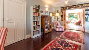 A spacious living room with hardwood flooring, a large bookshelf, a patterned armchair, and a dining area with a table and chairs. - Pass the Keys | Charming 3-Bed Bungalow with Character & Garden (London)