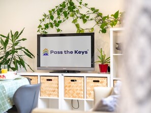 Un téléviseur affichant l’émission « Pass the Keys » sur un écran fixé au mur, entouré de plantes et d’un meuble blanc contenant des boîtes de rangement.