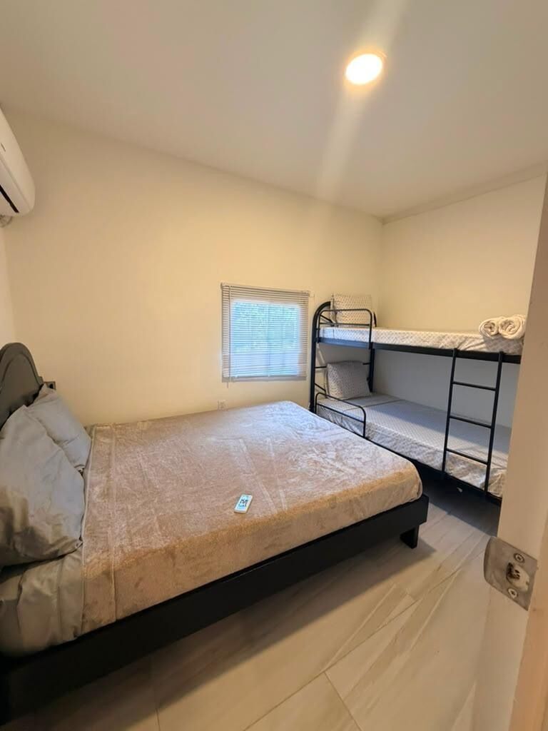 A small, single-room accommodation with a bunk bed, a single bed, a window with blinds, and a ceiling light.