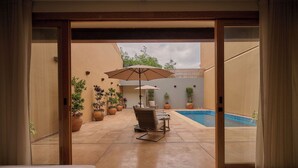 Una habitación con cama, puerta de vidrio corrediza que da a un patio con piscina, mobiliario exterior y plantas en macetas.