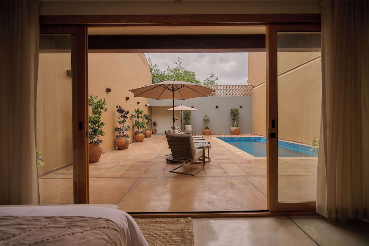 A room with a bed, a sliding glass door leading to a patio with a pool, outdoor furniture, and potted plants.
