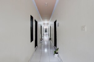 Um corredor comprido e estreito com paredes brancas e chão de azulejos, que leva a um quarto com janela.