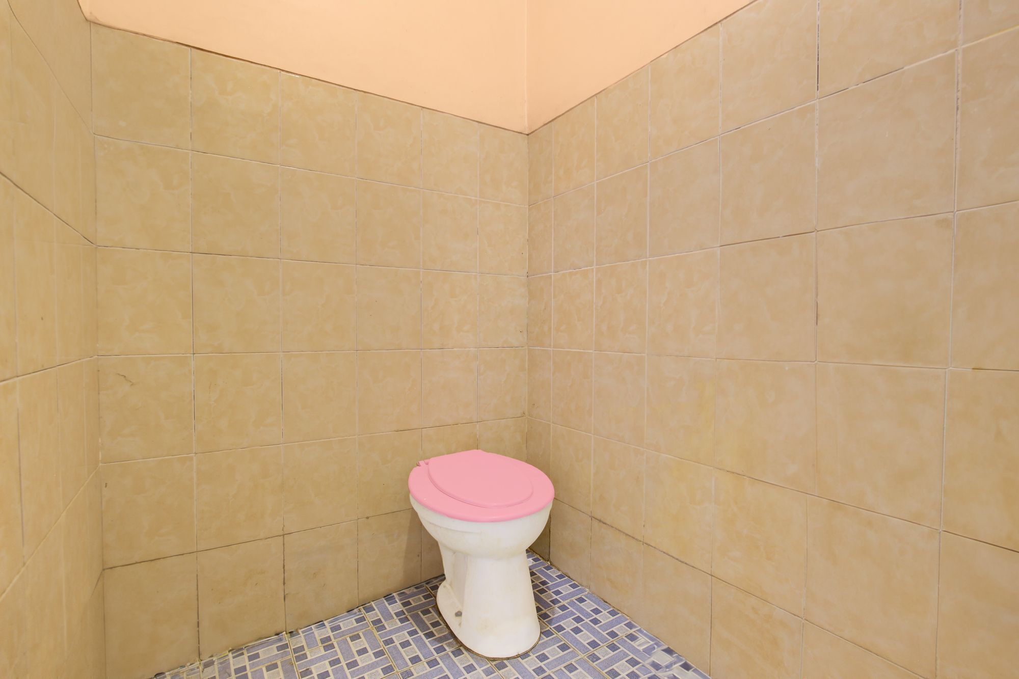 Een badkamer met beige betegelde wanden en vloer, een wit toilet met een roze zitting, en een vloer met een betegeld patroon.