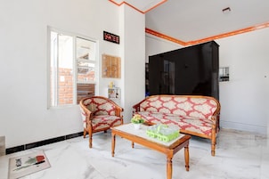Sala de estar com sofá, duas poltronas, uma mesa de centro e uma mesinha com garrafas. Há uma janela, uma porta e um relógio de parede.