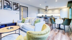 A modern living room with a sofa, coffee table, and chairs, a dining area with a table and chairs, and a kitchen with cabinets and a stove.
