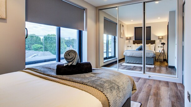 A modern bedroom with a large bed, a view of the outdoors through a sliding glass door, and a camera on a tripod in the corner.