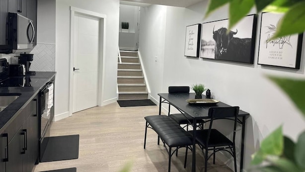 A modern kitchen with dark cabinetry and a black countertop, a dining area with a bench and chairs, and a staircase leading to an upper level.