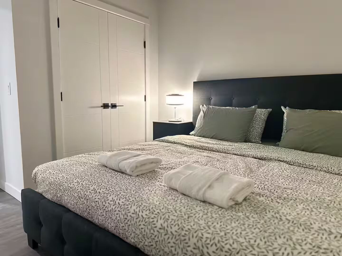 Een netjes opgemaakt bed met een beddengoed met patroon en twee opgevouwen handdoeken, een donker hoofdbord, een bedlampje en een witte, gesloten garderobe.