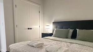 A neatly made bed with a patterned bedspread and two folded towels, a dark headboard, a bedside lamp, and a closed white wardrobe.