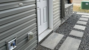 A house with a gray siding exterior, a white front door, a small porch with a stone ledge, and a green trash bin in the background.