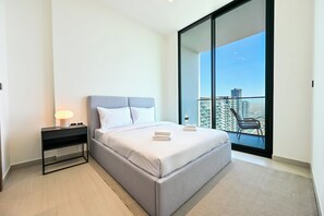 A modern bedroom with a large bed, a bedside table with a lamp, and a view of a cityscape through a large window. - Pure Voyage JVC (Dubai)