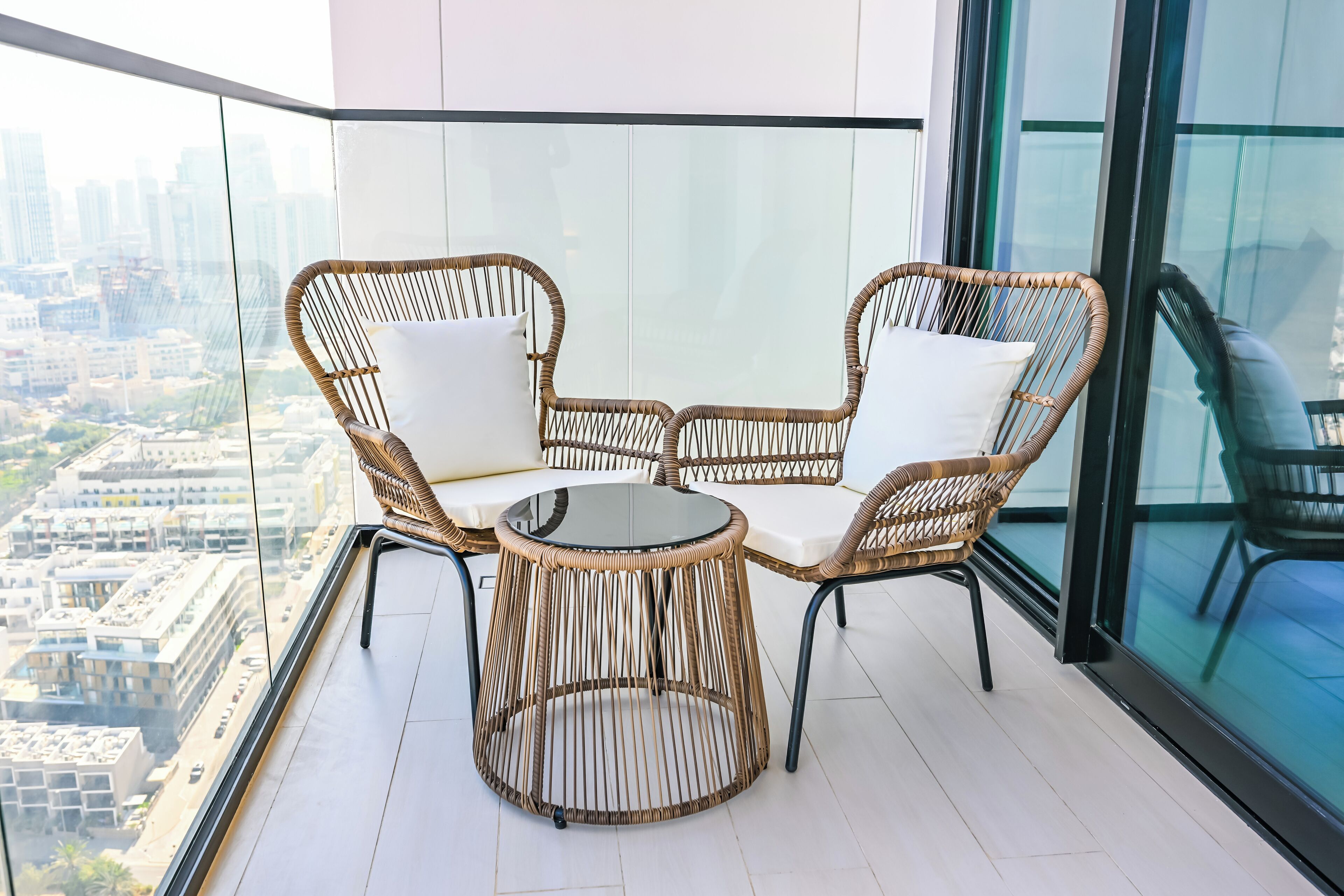 A balcony with wicker furniture and a glass table, overlooking a cityscape.