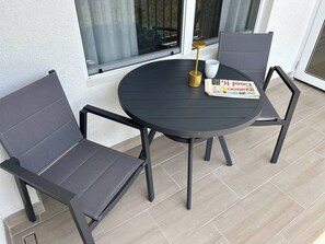 Een ronde tafel met twee stoelen, een krant en een kopje op een balkon met betegelde vloer en een raam op de achtergrond.