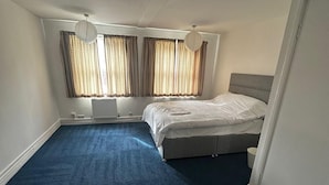 A bedroom with a bed, two windows with curtains, a radiator, and a blue carpet. - 262 Apartments (Leeds)