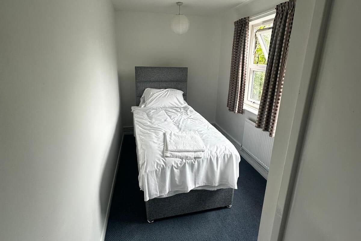 A narrow, single-bed room with a grey headboard, white bedding, a radiator, and a window with curtains.