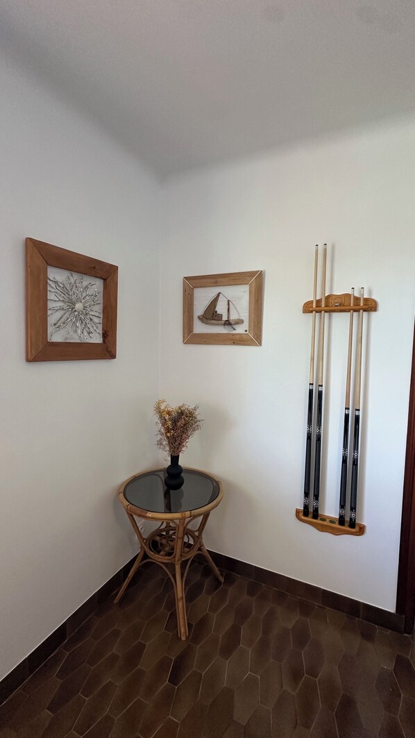 A corner room with a round glass table, two framed artworks, a vase with dried flowers, and a wall-mounted pool cue rack.