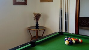 A pool table with a green felt surface, a white cue ball, and a set of pool cues. There are two framed artworks on the wall and a small table with a vase of dried flowers. - Sleeps 11, close to the sea (Tréogat)