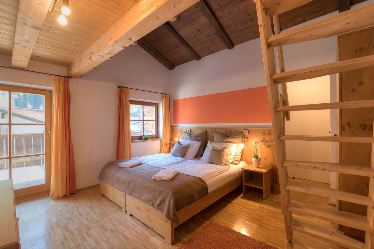 A bedroom with a wooden bed, a nightstand, a window with curtains, and a wooden ladder.
