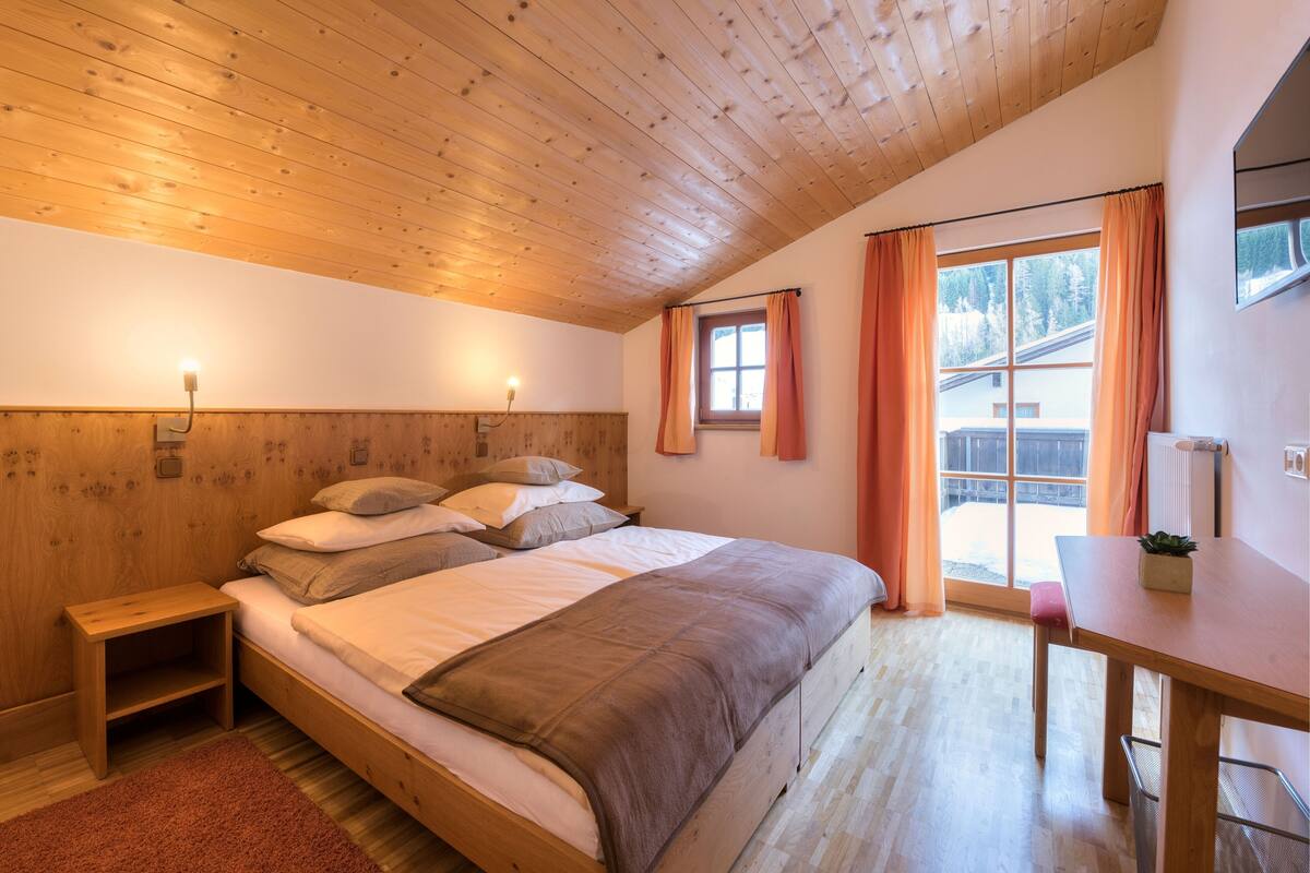 A bedroom with a wooden ceiling, a bed with pillows, a nightstand, a table, and a window with curtains.