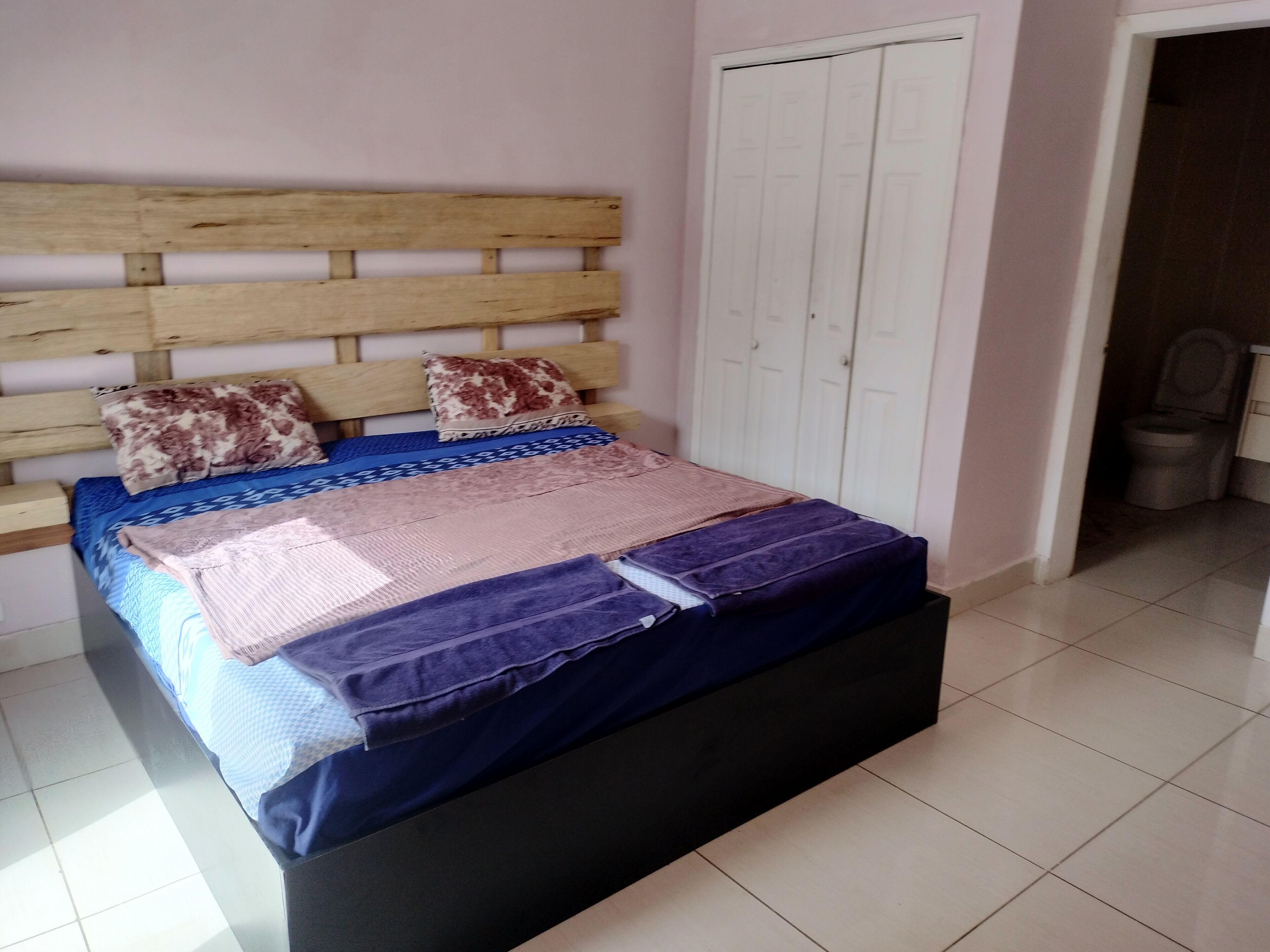 Ein Schlafzimmer mit hölzernem Kopfteil, einem Bett mit mehreren Decken, einem Kleiderschrank und einem angrenzenden Badezimmer mit Toilette.
