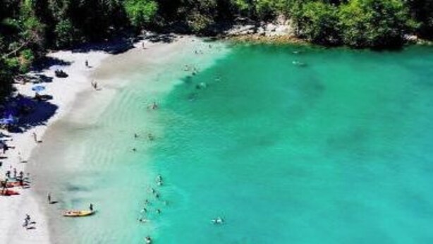 An aerial view of a beach with clear turquoise water, surrounded by dense greenery.