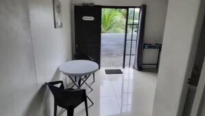 A narrow hallway with a small table and chairs, a door with a 'Welcome' sign, and a view of greenery outside.