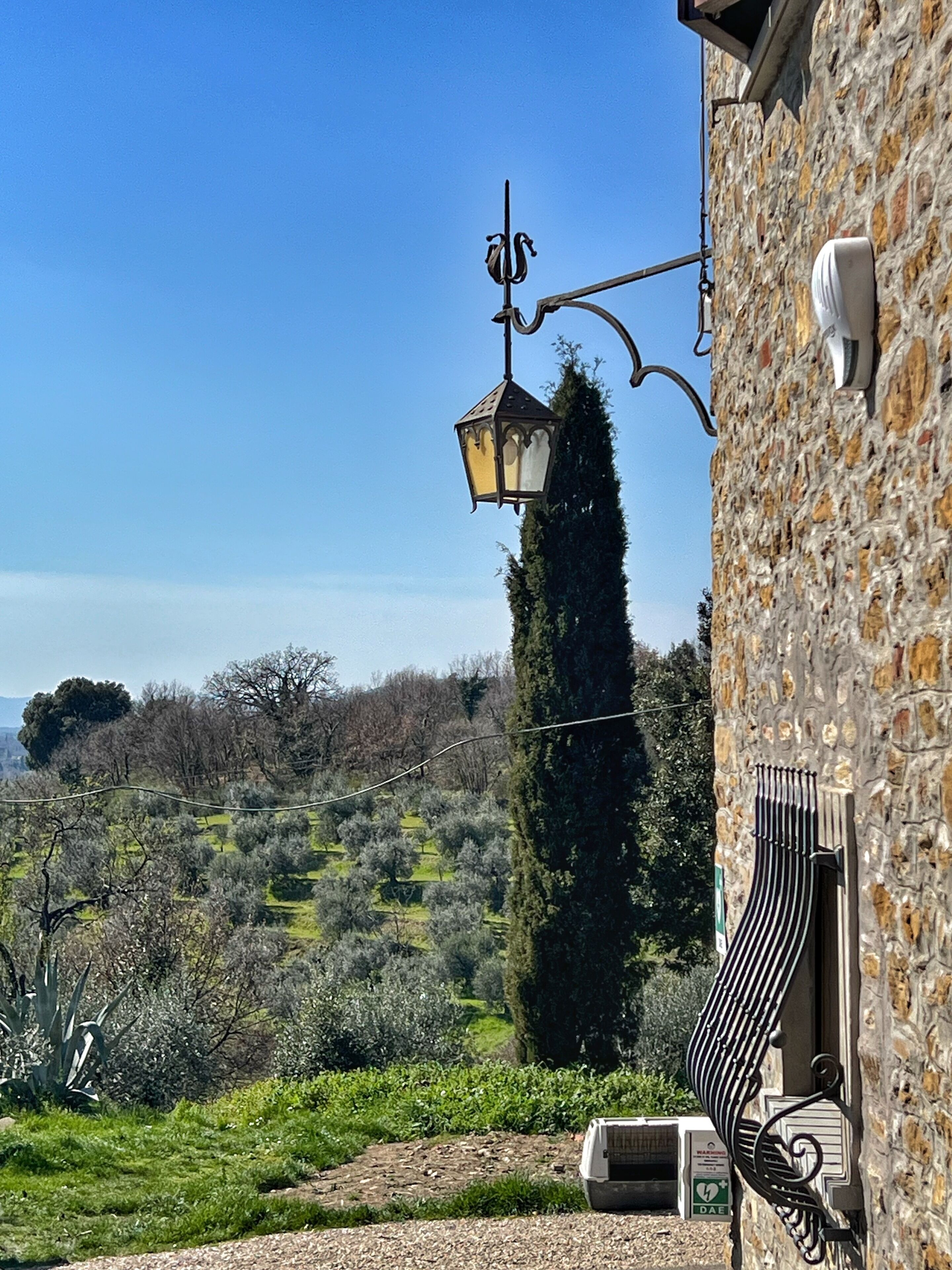 A stone building with a hanging lantern, a stone wall, and a view of a green landscape with trees and a hill in the background.