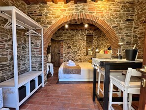 A rustic room with a stone wall, a wooden door, a bed with a white blanket, a wooden bench, a table with a bucket, and a mirror. - La Corte di Muciafora (Muciafora)