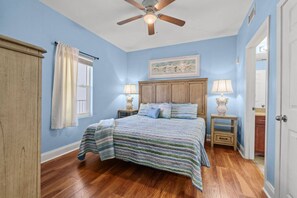 Une chambre à coucher comprenant un lit, deux tables de chevet avec des lampes, un ventilateur de plafond, une fenêtre avec des rideaux et une porte.