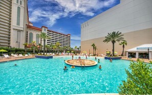 Una gran piscina al aire libre con varias pistas, rodeada de sillones de descanso y palmeras.
