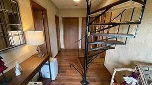 A staircase with a metal railing leading to a landing. There is a wooden table with a lamp and decorative items on the left, and a wicker chair with a cushion on the right. - Delightfully rustic 4-bedroom house in Vilas with stunning long-range views. (Vilas)