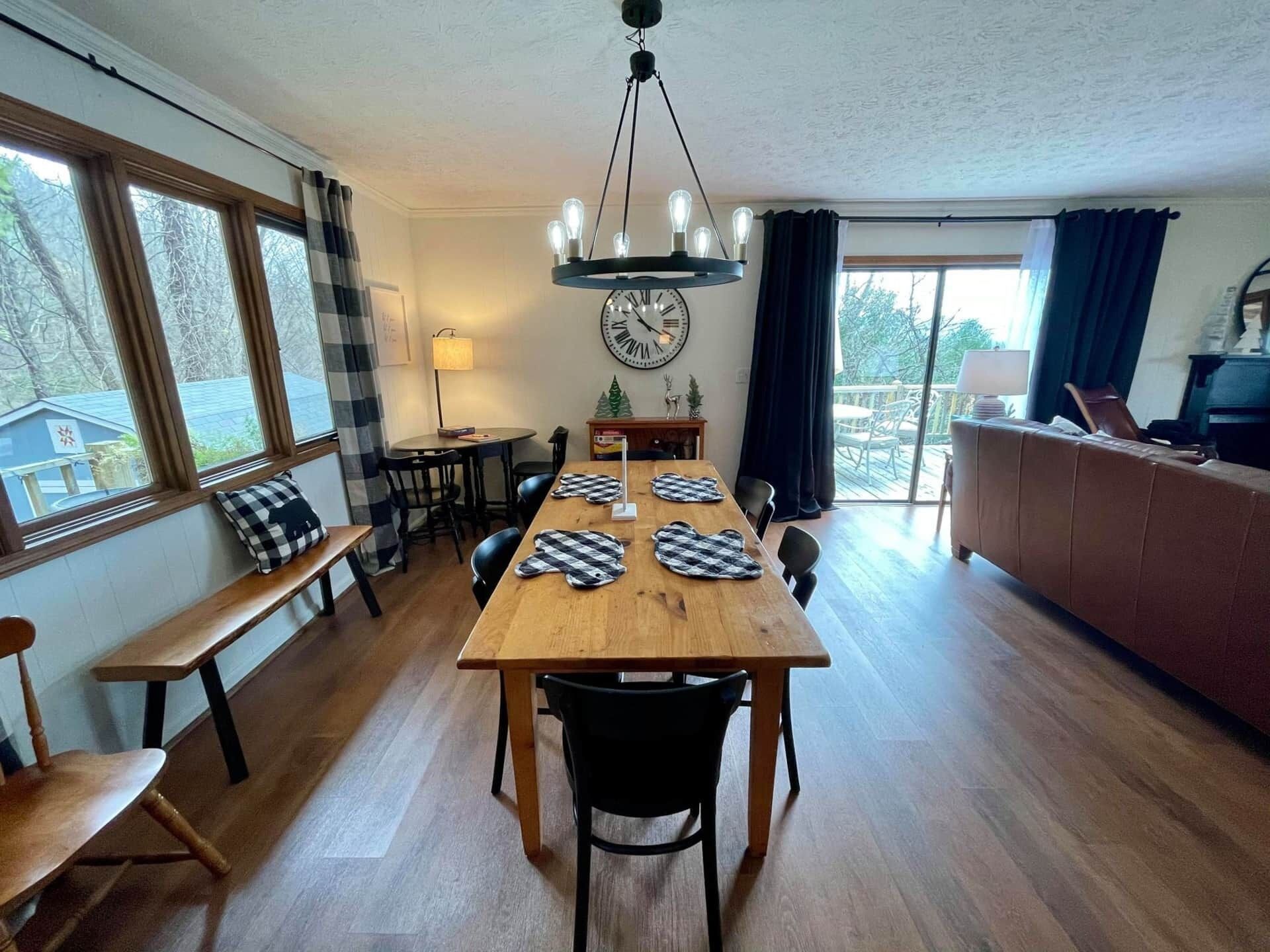 Uma sala de jantar com mesa de madeira, cadeiras pretas, um candelabro e vista para o exterior através de grandes janelas.
