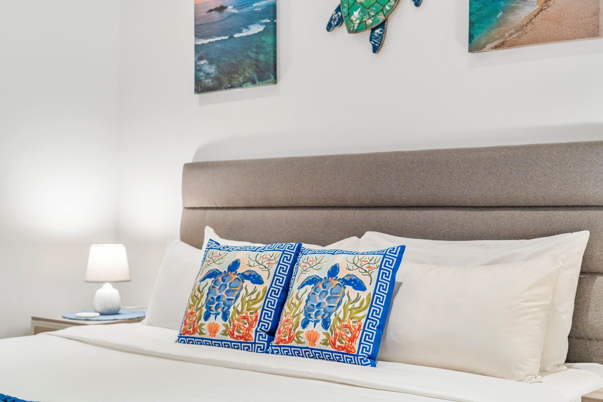 Een keurig opgemaakt bed met twee decoratieve kussens, een bedlampje en een wand met ingelijste kunstwerken.