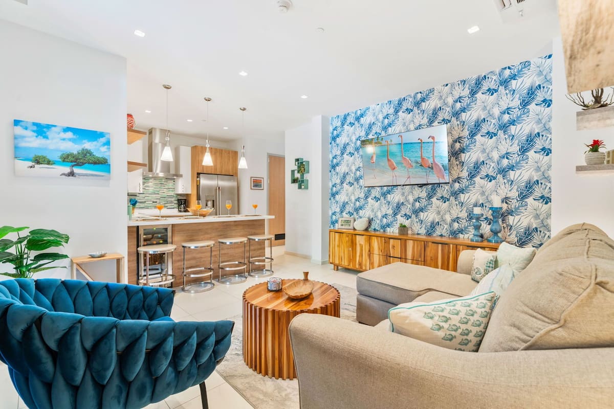 A modern living room with a sofa, a blue armchair, a wooden coffee table, and a kitchen area in the background.