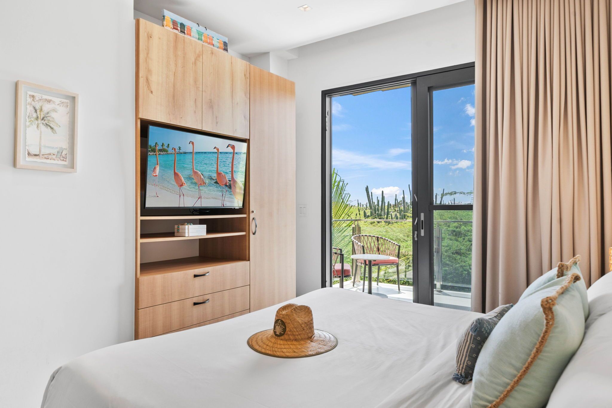 A bedroom with a bed, a TV on a wooden cabinet, a balcony with a chair, and a view of the ocean.