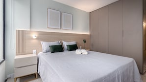 Ein ordentlich bezogenes Bett mit weißer Bettdecke, zwei Kissen und einem Paar gefalteter Handtücher. Ein Nachttisch mit Lampe steht daneben, und der Raum zeichnet sich durch ein modernes Design, eine helle Wand und ein Holz-Kopfteil aus.