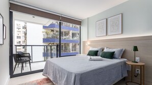 Ein modernes Schlafzimmer mit einem großen Bett, einem kleinen Nachttisch, einem Sessel und einem Balkon mit Blick auf ein blau gefliestes Gebäude.