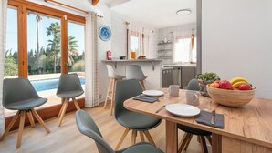 A dining area with a wooden table, grey chairs, a bowl of fruit, and a view of a swimming pool. - Villa Zahara - paceful retreat in the countryside (Alcúdia)
