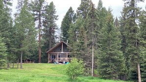 A cabin with a deck, surrounded by tall trees and a pond.