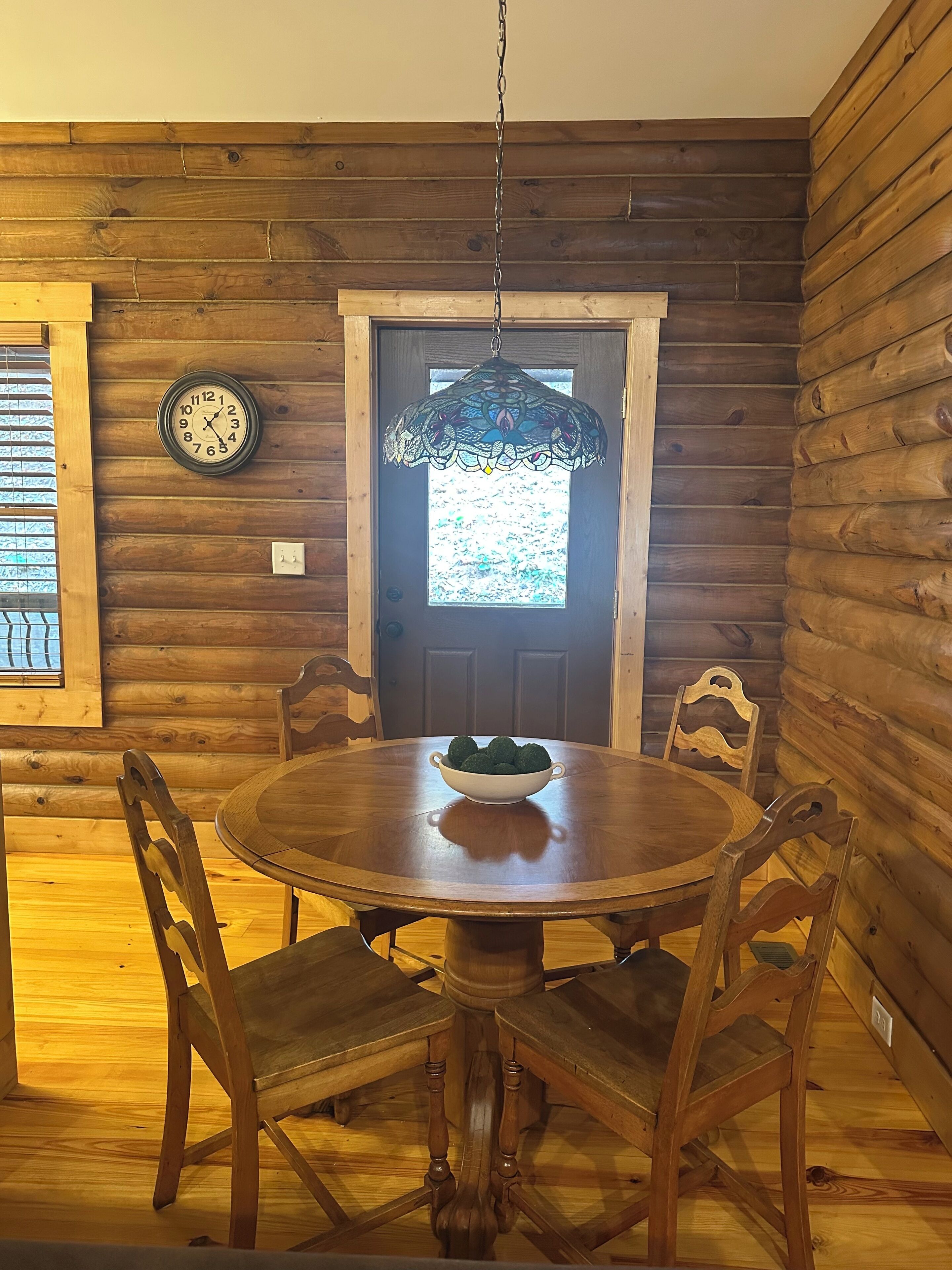 A wooden interior with a round dining table, four chairs, a wall clock, and a decorative hanging light fixture.