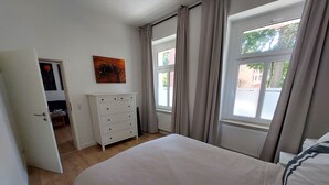 Un dormitori amb un llit, una petita taula de nit, un quadre a la paret i una finestra amb cortines.